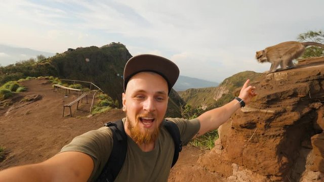 Young Bearded Blogger Man Taking Selfie Video While Hiking on the Top of Mountain and Meet Wild Monkey. 4K Lifestyle Travel Concept Footage. Batur Volcano, Bali, Indonesia.