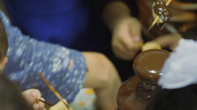Chocolate fountain at a children's party. Dipping fruit pieces on wooden sticks in a brown dessert.