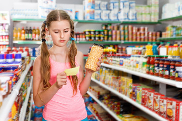 Positive tween girl choosing food products on shopping list in supermarket