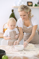 Obraz na płótnie Canvas Little girl and her blonde mom in beige aprons playing and laughing while kneading the dough in kitchen. Homemade pastry for bread, pizza or bake cookies. Family fun and cooking concept