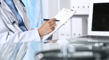 Female doctor filling up medical form on clipboard, closeup. Reflecting glass table is a physician...