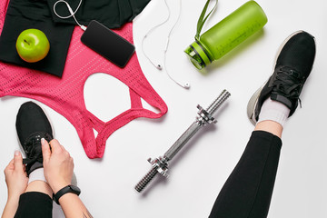 Young woman in sporting leggings laces sneakers, preparing for training. Accessories for sports on white background flat lay top view.