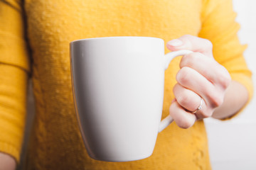Female hand holding white cup in yellow sweater background. Copy space