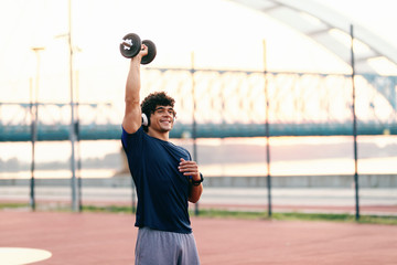 Sporty muscular man with curly hair lifting dumbbell while standing on the court in the morning. Headphones on ears.