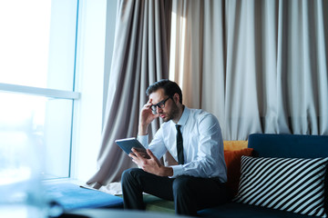 Pensive Caucasian businessman using tablet and holding head while sitting on sofa in office.