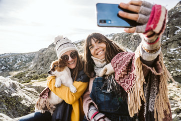 Fototapeta premium .Two young women, covered with a blanket, on a sunny winter morning enjoying a day on the mountain with their cute little dog taking themselves pictures with their smartphone. Lifestyle.