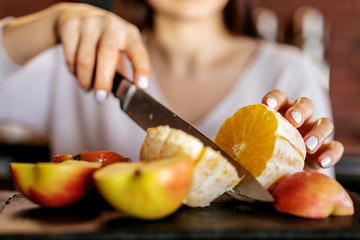 A young woman-cook slicing fruits: apple, kiwi and orange, preparing to make juice with electric juicer.
