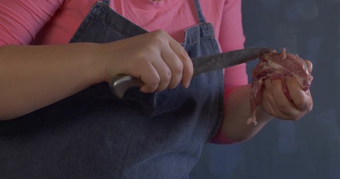 Slim woman cook in pink sweater, grey apron and white cap splits the leg of pig on wooden board. Grinds small pieces of meat with meat grinder and sorts minced meat into packages.