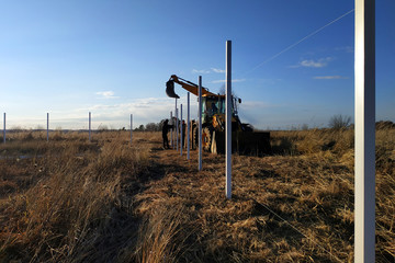 Fototapeta na wymiar The excavator with the bucket clogs the iron pillars for the fence in the field, the pillars painted with white paint