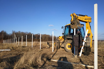 The excavator with the bucket clogs the iron pillars for the fence in the field, the pillars painted with white paint