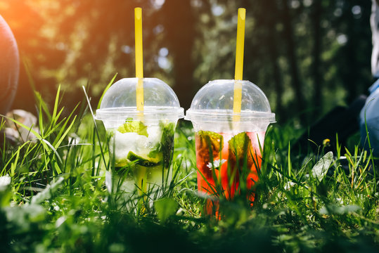 Two Summer Cocktails Standing Outdoors In The Green Grass. Cold Non-alcoholic Drinks With Ice To Go. Mojito And Strawberry Lemonade With Lime, Soda Water And Mint. Freshness And Holidays.