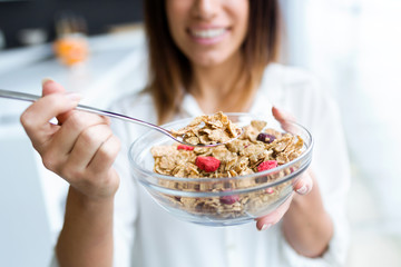Pretty young woman showing cereals to camera while eating in the kitchen at home.