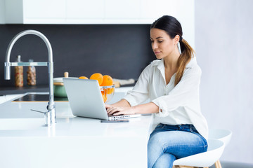 Pretty young woman using her laptop in the kitchen at home.