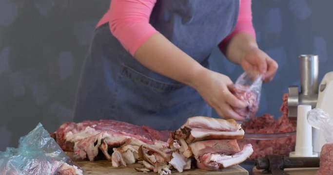 Slim woman cook in pink sweater, grey apron and white cap splits the leg of pig on wooden board. Grinds small pieces of meat with meat grinder and sorts minced meat into packages.