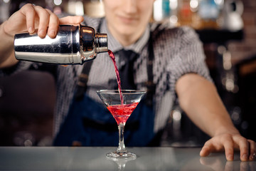Expert bartender making cocktail with shaker in martini glass, red. Dark background