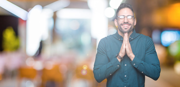 Young handsome business man wearing glasses over isolated background praying with hands together asking for forgiveness smiling confident.