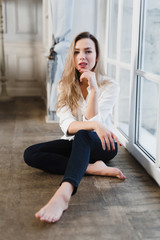 Fototapeta na wymiar Model blonde white shirt blue pants jeans sitting on wooden parquet floor by window.