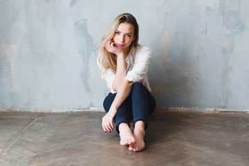 Fototapeta premium Model blonde barefoot blue jeans white shirt sitting on floor against gray wall.