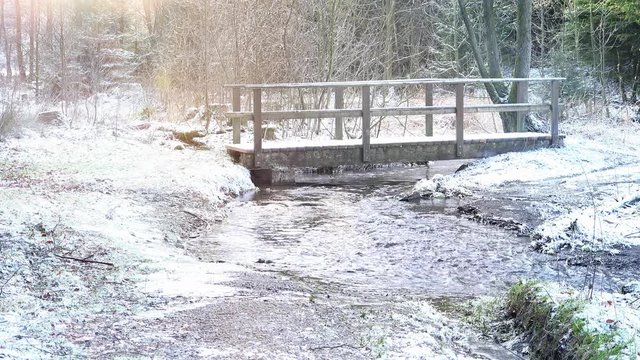 Im Winterwald flie&szlig;t ein Flu&szlig;. Es ist die Glenne bei Rixen im Sauerland. Eine Holzbr&uuml;cke &uuml;berquert den Fluss. Sound. Die Naturlandschaft ist mit Schnee bedeckt.
