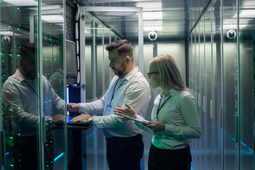 Diverse adult man and woman using tablet and laptop while diagnosing server hardware in modern data center
