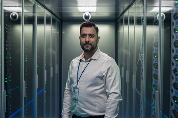 Adult IT worker in white shirt standing among server racks in data center smiling at camera