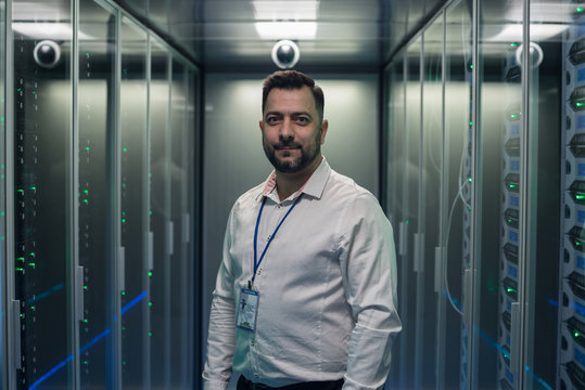 Adult IT Worker In White Shirt Standing Among Server Racks In Data Center Smiling At Camera