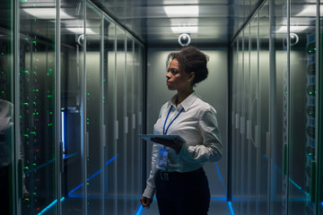 African American woman using tablet while walking in corridor of data center and checking hardware...