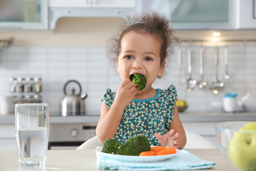 Cute African-American girl eating vegetables at table in kitchen