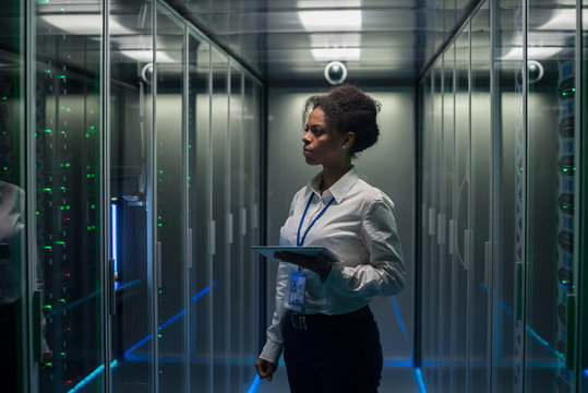 African American woman using tablet while walking in corridor of data center and checking hardware on server racks