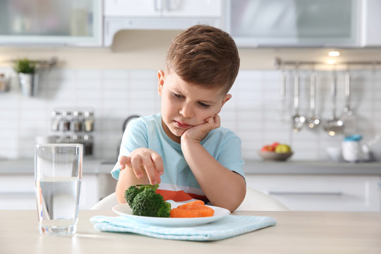 Unhappy Little Boy Eating Vegetables At Table In Kitchen