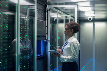 Medium shot of female technician working on a tablet in a data center full of rack servers running diagnostics and maintenance on the system - obrazy, fototapety, plakaty