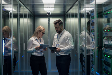 Side view of man and woman with tablet diagnosing server hardware opening glass door of rack in data center corridor