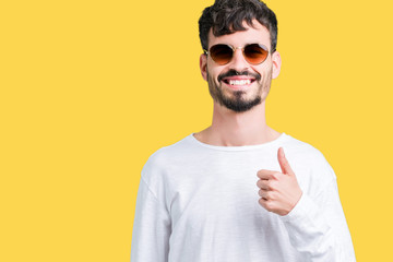 Young handsome man wearing sunglasses over isolated background doing happy thumbs up gesture with hand. Approving expression looking at the camera with showing success.