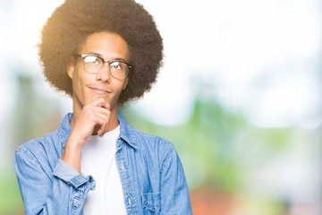 Young african american man with afro hair wearing glasses with hand on chin thinking about question, pensive expression. Smiling with thoughtful face. Doubt concept.