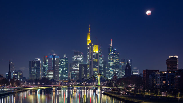 Lunar eclipse at the end of totality over the skyline of Frankfurt, Germany, with reflections on the Main.