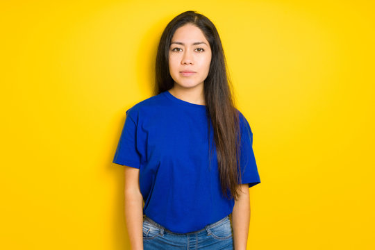 Beautiful brunette woman wearing blue t-shirt over yellow isolated background with serious expression on face. Simple and natural looking at the camera.
