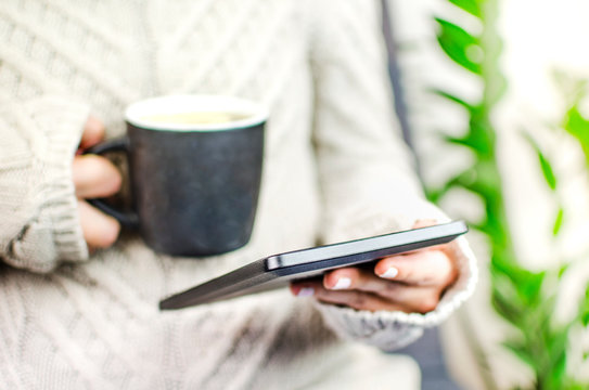 Female person wearing sweater holding electronic reader and a hot beverage