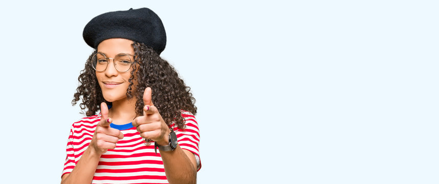 Young beautiful woman with curly hair wearing glasses and fashion beret pointing fingers to camera with happy and funny face. Good energy and vibes.