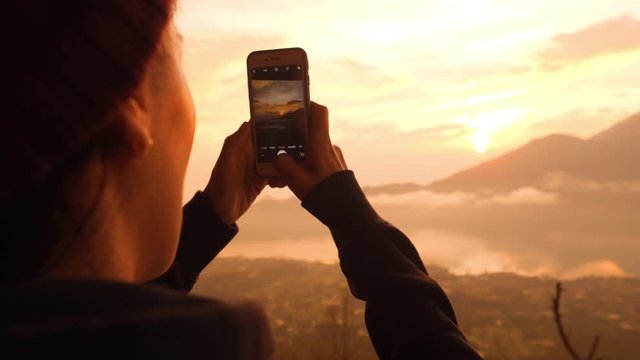 Young Mixed Race Tourist Hipster Girl Taking Photos of Amazing Sunrise from Top of Batur Volcano Mountain Using Mobile Phone. 4K Slowmotion Travel and Freedom Lifestyle Concept Shot. Bali, Indonesia.