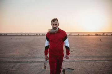 Handsome bearded mature man in sports suit walking on beach, outdoors during sunset or sunrise