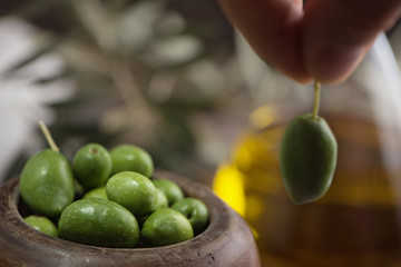 Wooden cup of olives and fingers with an olive on olive oil in glass bottle background.