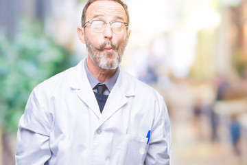 Middle age senior hoary professional man wearing white coat over isolated background making fish face with lips, crazy and comical gesture. Funny expression.