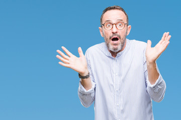 Fototapeta premium Middle age hoary senior man wearing glasses over isolated background clueless and confused expression with arms and hands raised. Doubt concept.