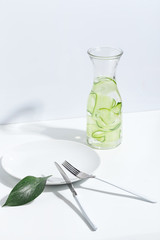 Plate, cutlery and jar of cucumber water on a white table. Creative minimalistic concept.