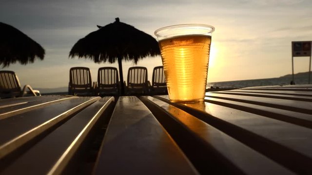 Cold beer on beach reveals setting sun over ocean view. SLIDER