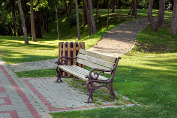 a bench with a wooden seat and iron forged legs with a wooden trash bin on paving slabs with a...