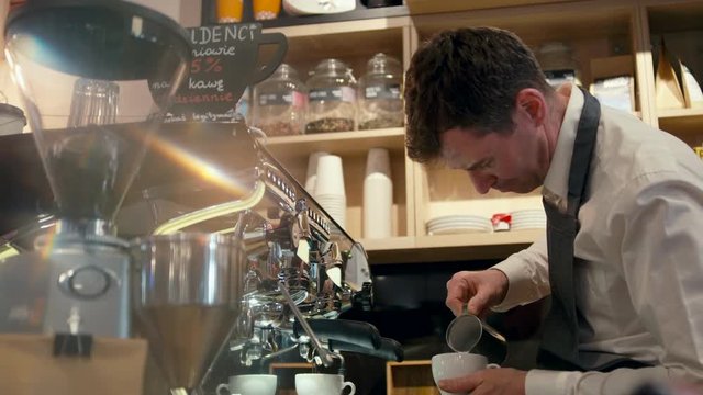 Barista Man in Apron is Making Cappuccino with Milk in Ceramic Cup using Professional Coffee Machine in the Coffee Shop or Cafe Bar