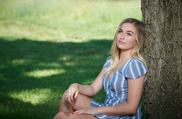 Stunning young blonde woman in summer dress sitting against tree in shade