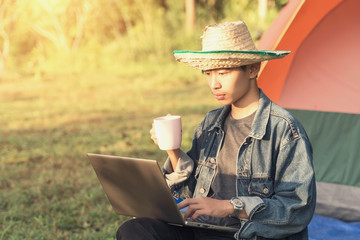 A young male student uses laptop in front of camping ten, holds cup of coffee in the morning on vacation holiday - Powered by Adobe
