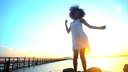 Young attractive African American woman with afro hair wearing Boho style sundress enjoying sunset dancing on the beach